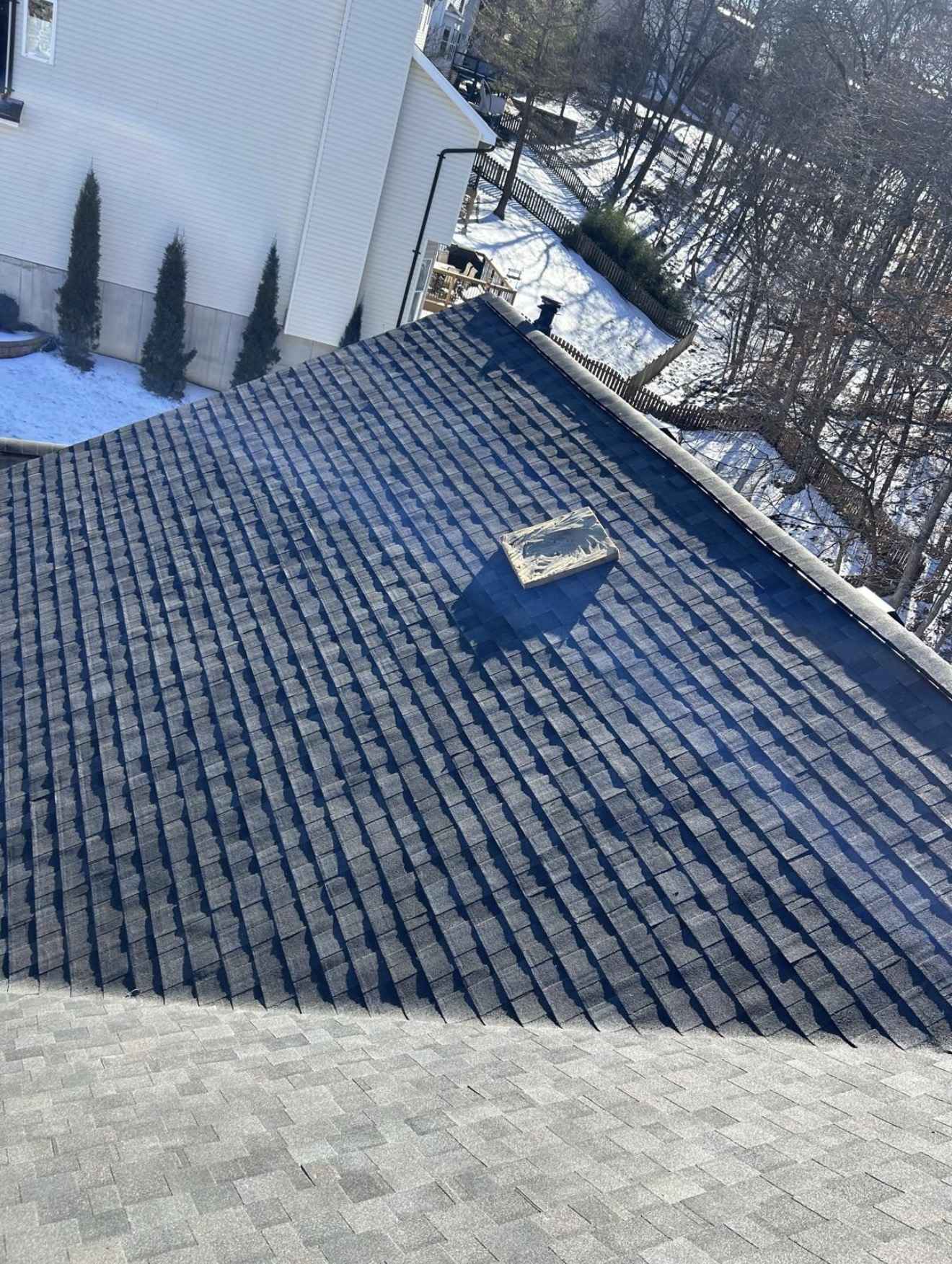 Roof mid installation photographed from the slope: courses of dark charcoal architectural shingle laid across a large hip plane, meeting an older lighter grey shingle surface at the lower hip in the foreground, with an offcut of new shingle lying loose, a cream lap sided wall and gutter above, a rear deck, a black metal fence and snow lying on a wooded slope behind