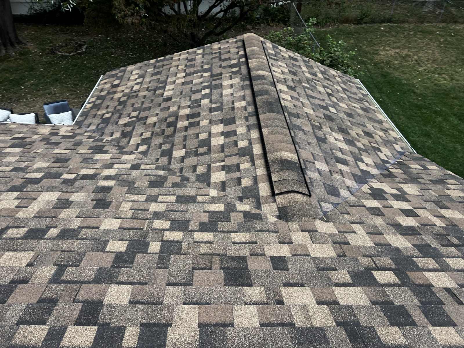 Looking down onto the hip of a newly laid brown, tan and charcoal multi tone architectural shingle roof, capped along the hip with ridge cap shingles and carrying a dark ridge vent strip, with the two roof planes falling away either side, a gutter along the eave, and a mown lawn, a chain link fence, a bare branched tree and cushioned garden furniture below