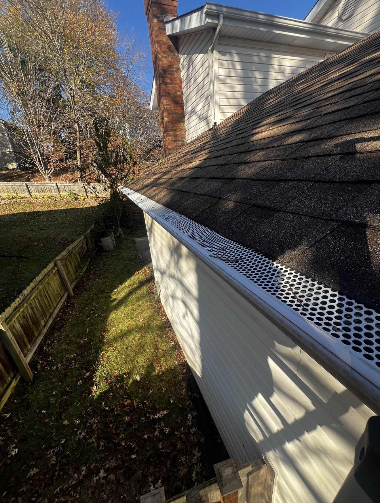 Looking along the eave of a weathered dark brown shingle roof on a white vinyl sided two storey house with a red brick chimney, where a white perforated metal gutter guard punched with round holes runs the length of the gutter with two bare twigs and a few small dark scraps of debris lying on top of the perforations, and a garden of mown grass, fallen autumn leaves and a wooden boundary fence below