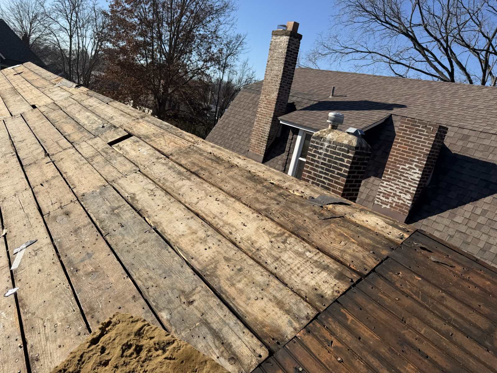 A roof plane stripped back to wide wooden plank decking, boards weathered and studded with old nail holes and with visible gaps between them, running up the left of the frame under a clear blue winter sky, with the main brick house behind still carrying its weathered brown shingles, two brick chimneys showing spalling and patched mortar, and a small dormer with a metal turbine vent