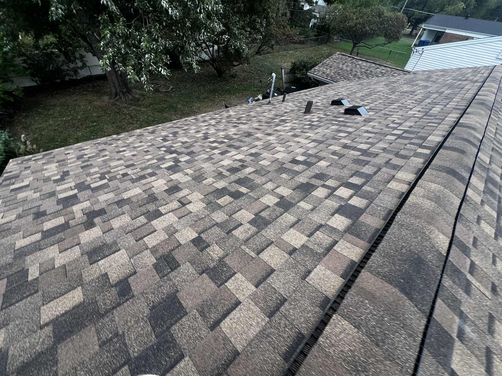 Wide view along a newly laid brown, tan and charcoal multi tone architectural shingle roof, with the capped ridge and a dark ridge vent running down the right hand side of the frame and two black box vents and a metal flue pipe set near the far end of the ridge, looking out over a back garden with a large tree, a chain link fence and a neighbouring roof