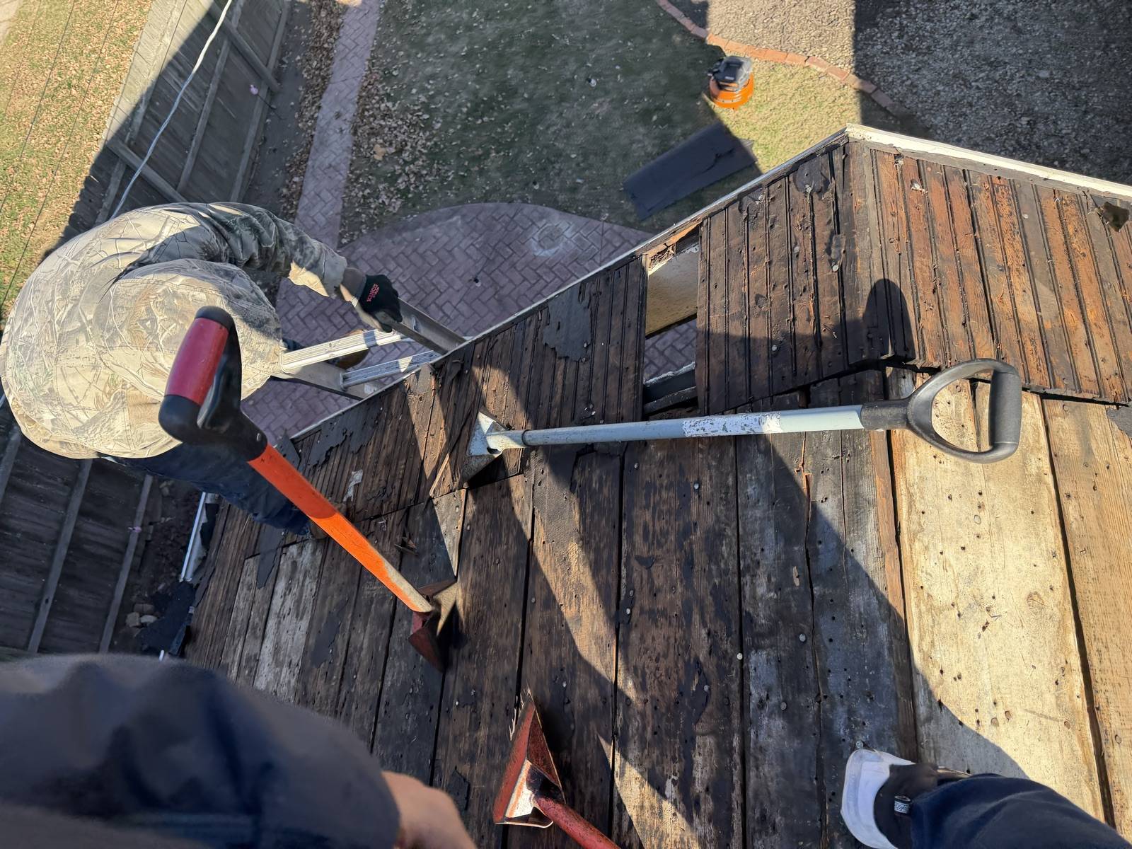 Looking down from a roof onto plank decking stripped bare of shingle, with an orange handled tear off bar, an aluminium D handle roofing shovel and two red flat spades lying across the boards, an aluminium ladder hooked over the eave, a crew member in a camouflage jacket and gloves crouched at the roof edge, and a brick paver patio, lawn and wooden fence in the garden below